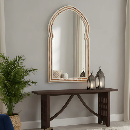 Decorative mirror with wooden frame on a wall above a dark wooden console table with lanterns and a plant.