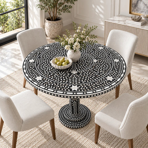 A round black and white patterned mother of pearl dining table with a pedestal base, surrounded by four cream upholstered chairs in a bright, modern dining room.