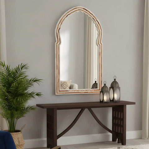 Decorative mirror with wooden frame on a wall above a dark wooden console table with lanterns and a plant.