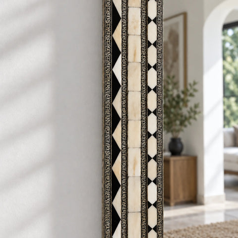 A tall, narrow mirror with an intricate black and cream bone-inlay geometric border, shown leaning against a white wall in a bright, modern room.
